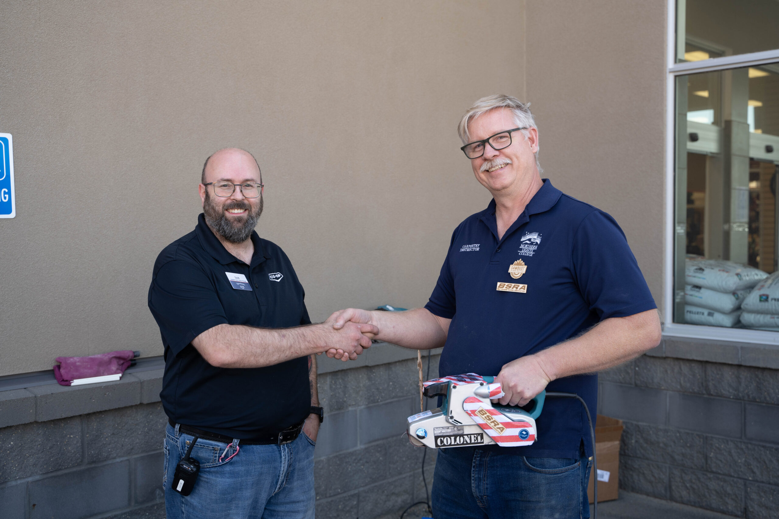 Co-op manager shakes hands with NLC instructor who is holding a belt sander