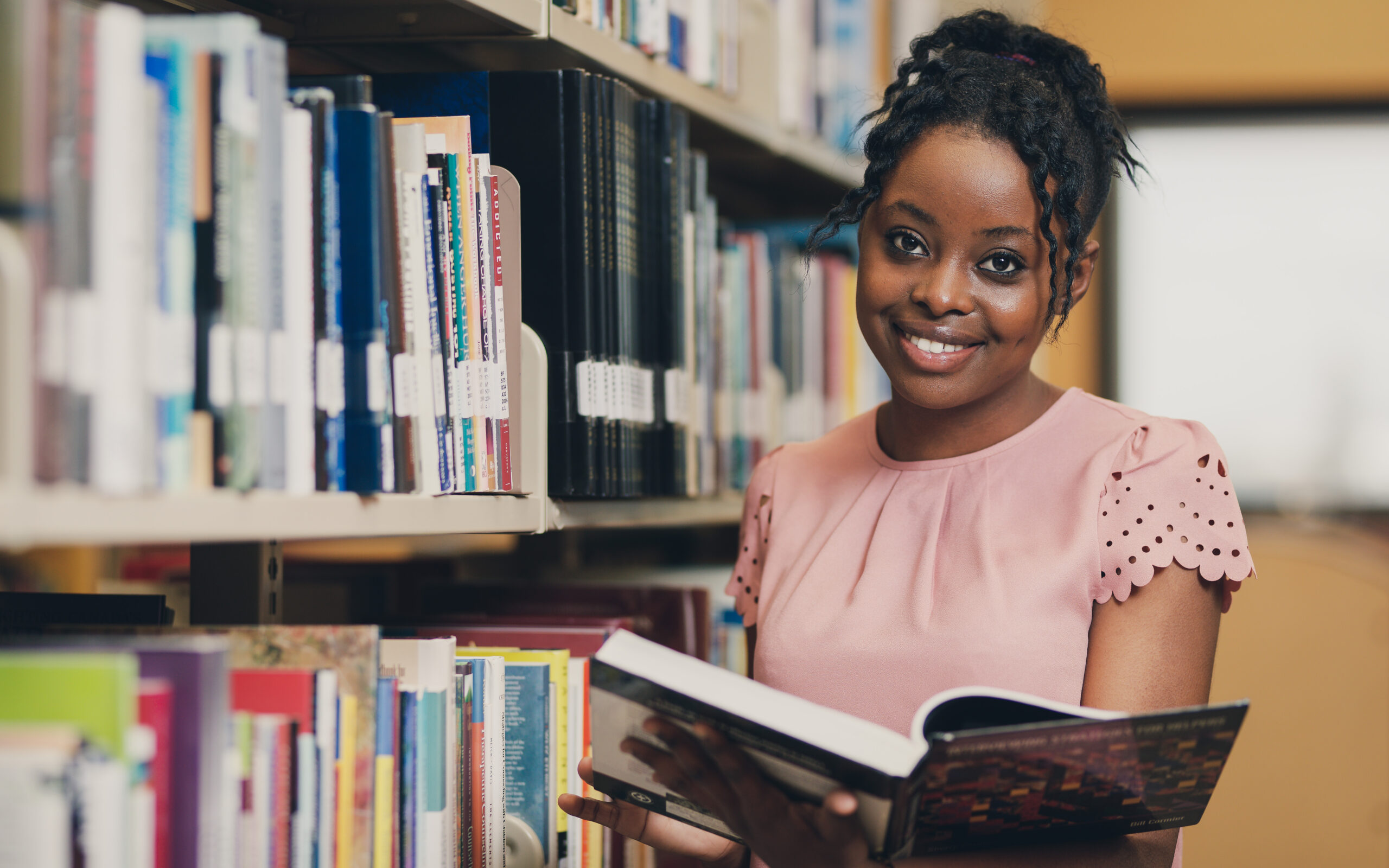 NLC Student in Library with book open and smiling at the camera